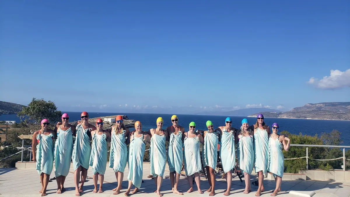 A group of 14 women wearing towels and swim caps pose for a photo in front of an ocean.