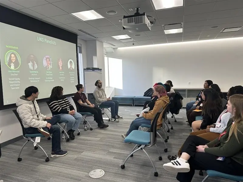 Four panelists sit in chairs in front of a screen at the front of a room, talking to college students.