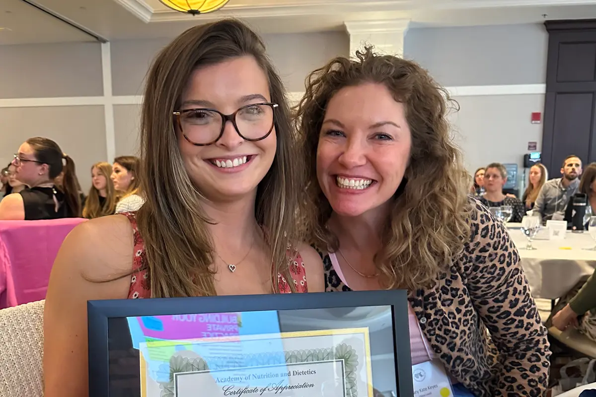 Two women smile for a photo. One of them is wearing glasses and holding an award.