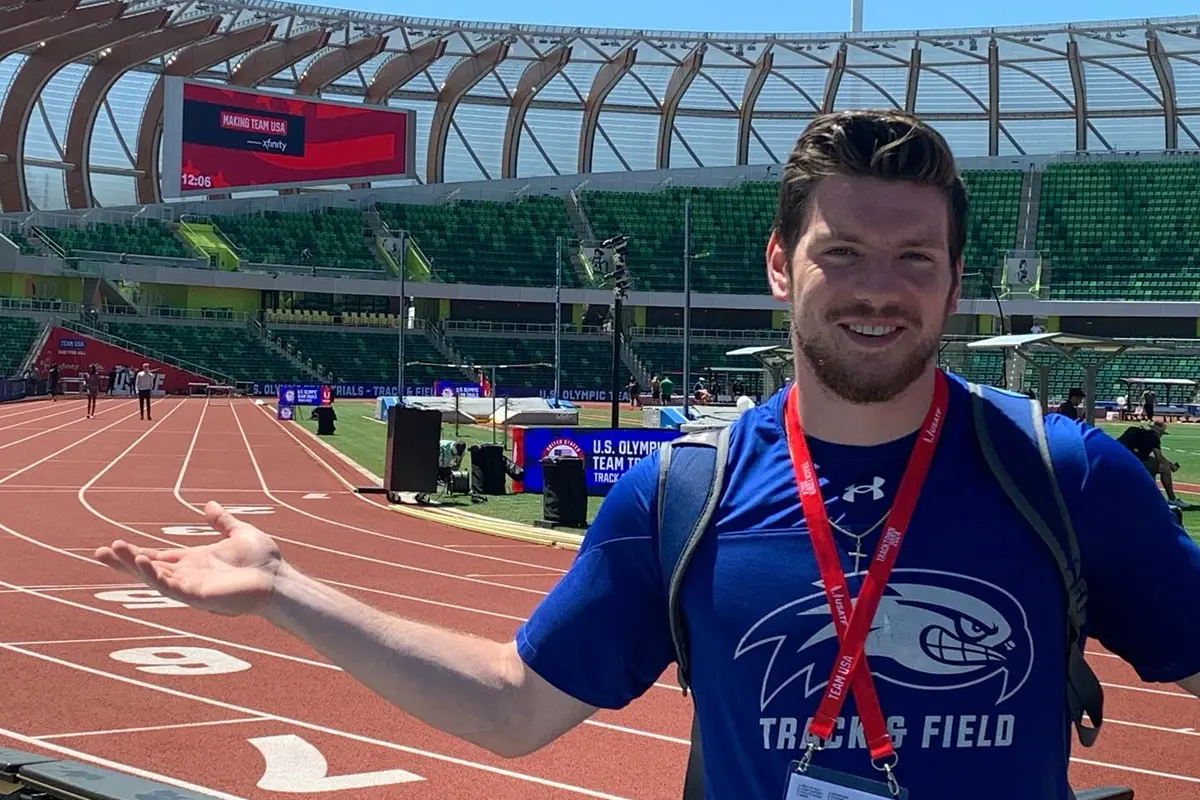 A person in a blue shirt gestures with his hand while posing for a photo at an outdoor track.