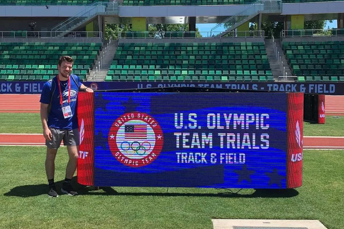 A person in a blue t-shirt poses for a sign for a track and field meet.