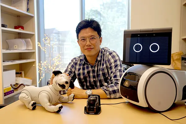 A man in glasses and a plaid shirt sits at a desk that has three robotos on it