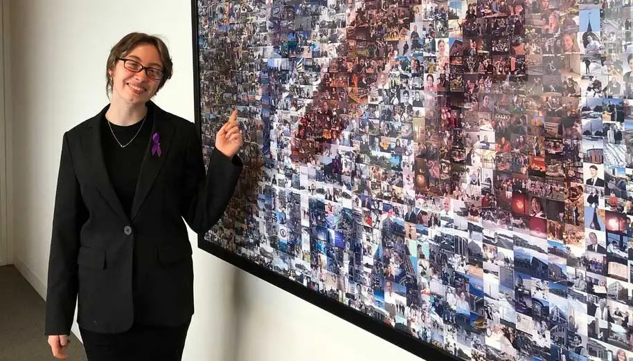 Meghan Dearing stands in front of a mosaic of small photos that are arranged to form the UMass logo.