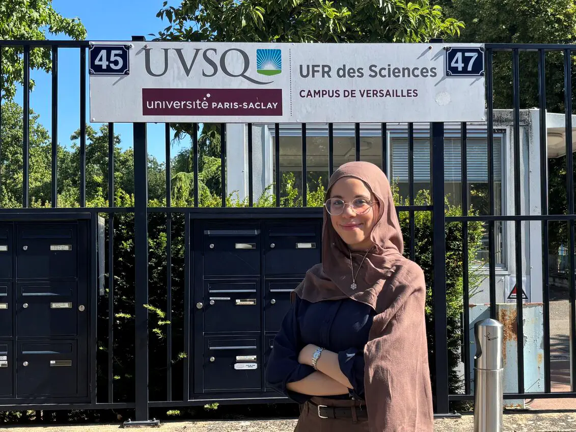A woman with glasses folds her arms and poses for a photo in front of a fence with a sign.