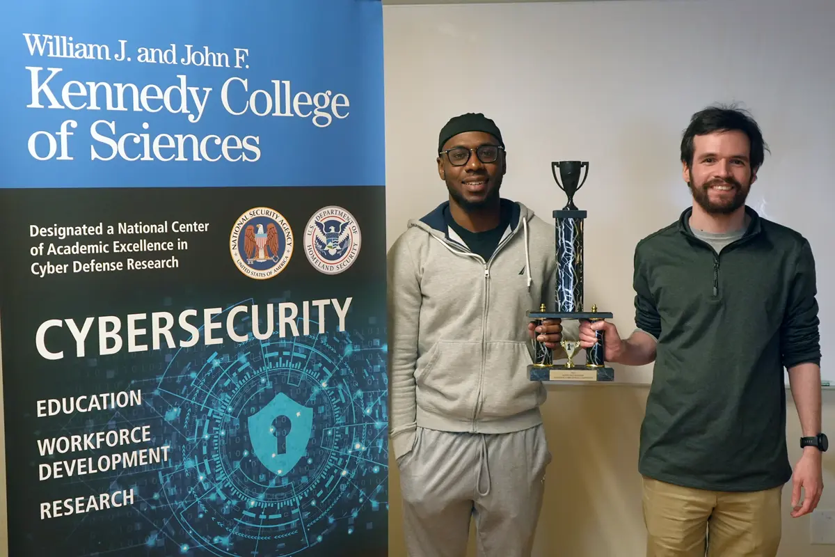 Two students hold a trophy next to a Kennedy College of Sciences Cybersecurity banner.