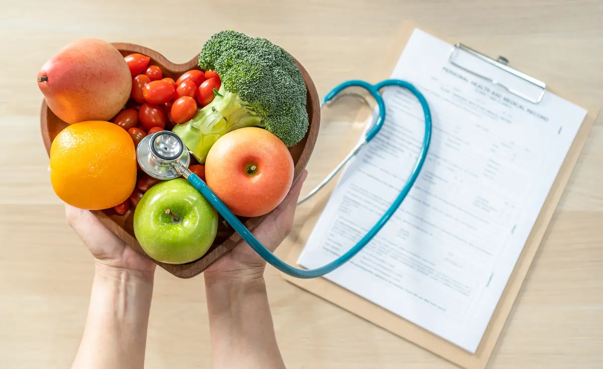 Fruits and vegetables in heart dish with stethoscope held above a nutritionist clipboard on table.
