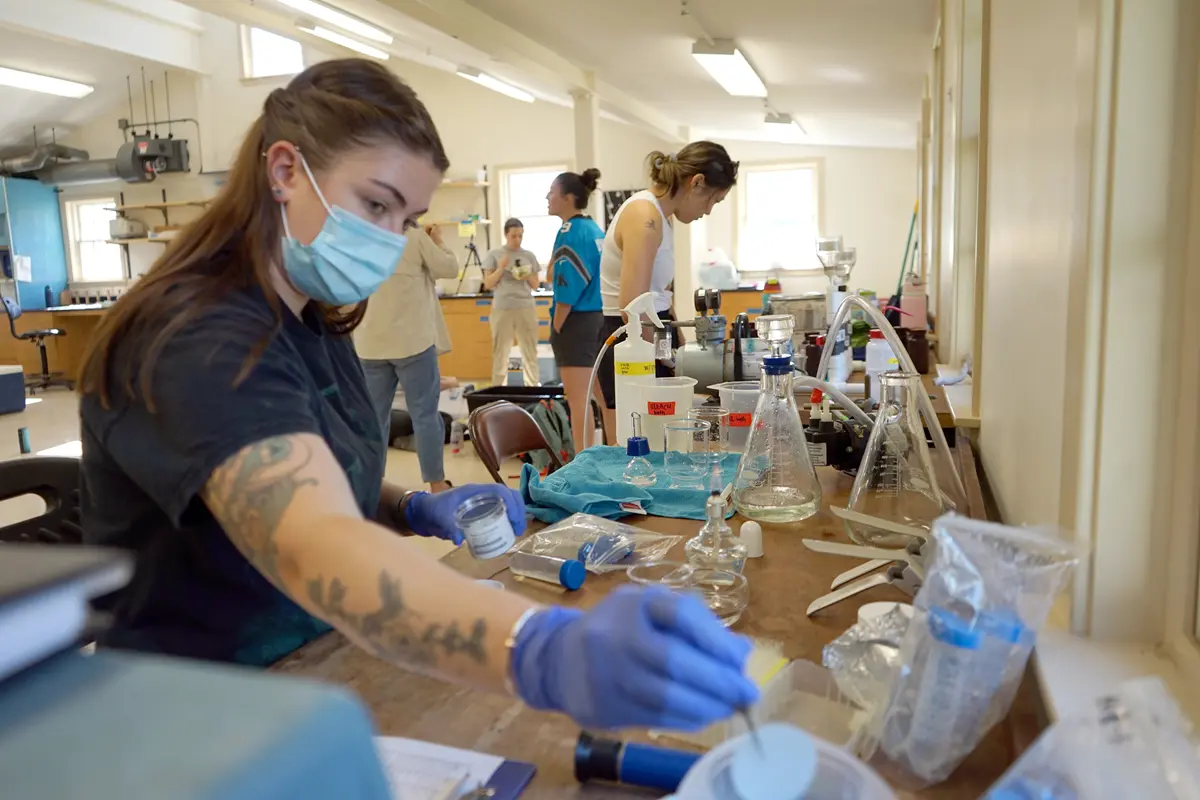 A young woman in a mask reaches for an item while working in a science lab.