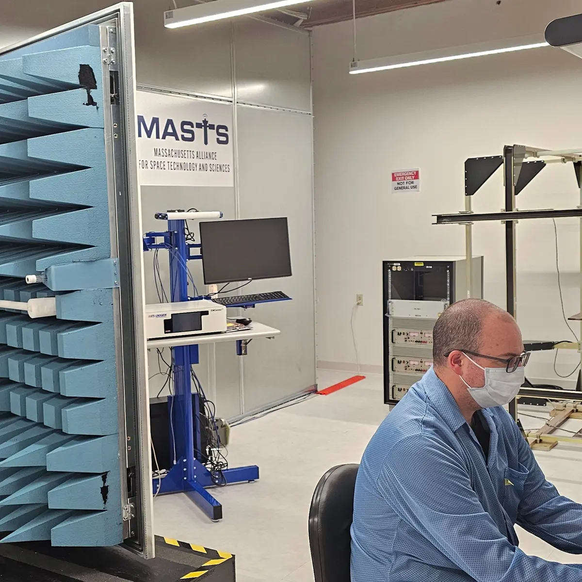 Man wearing a mask works at a laptop in front of metal door with blue spikes on it and MASTS sign on wall