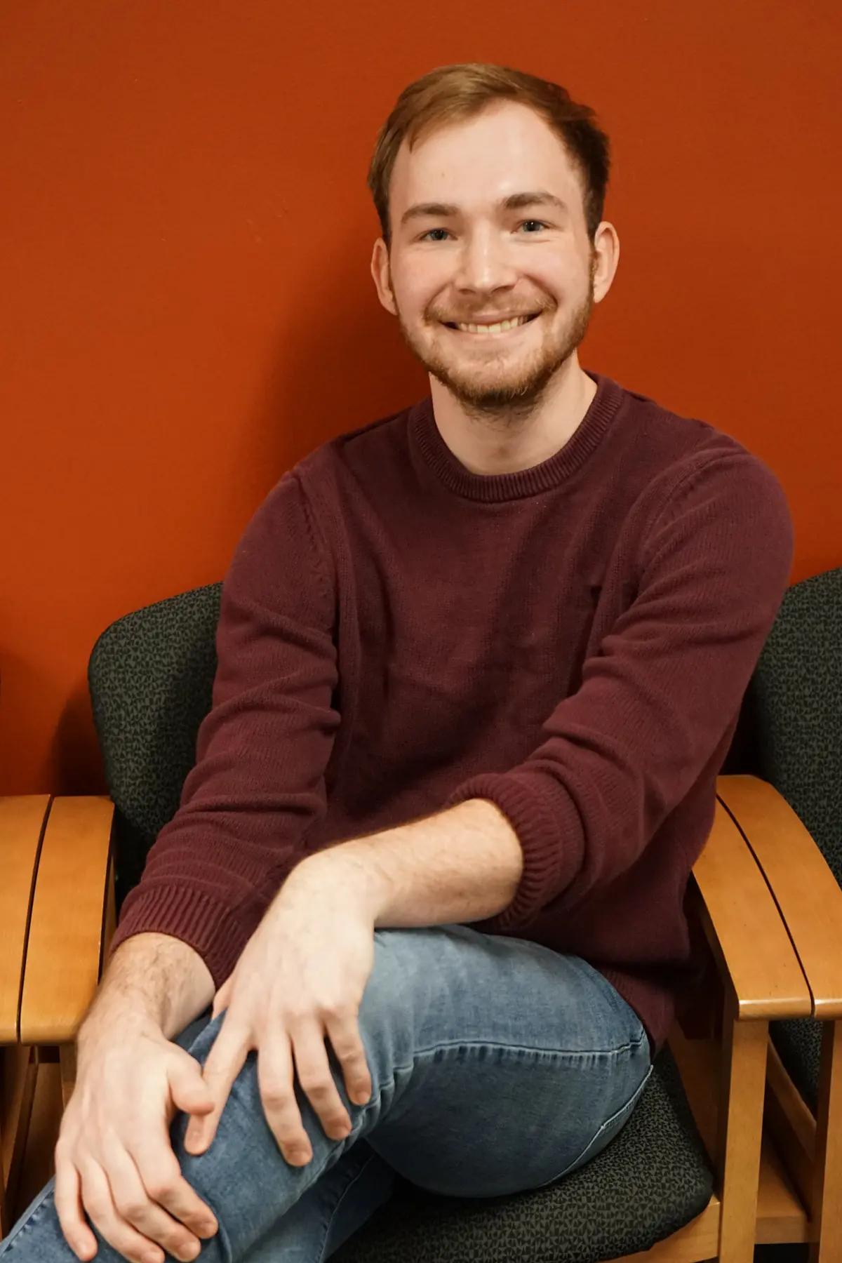 A young man with short hair crosses his leg while sitting in a chair and smiling for a photo.