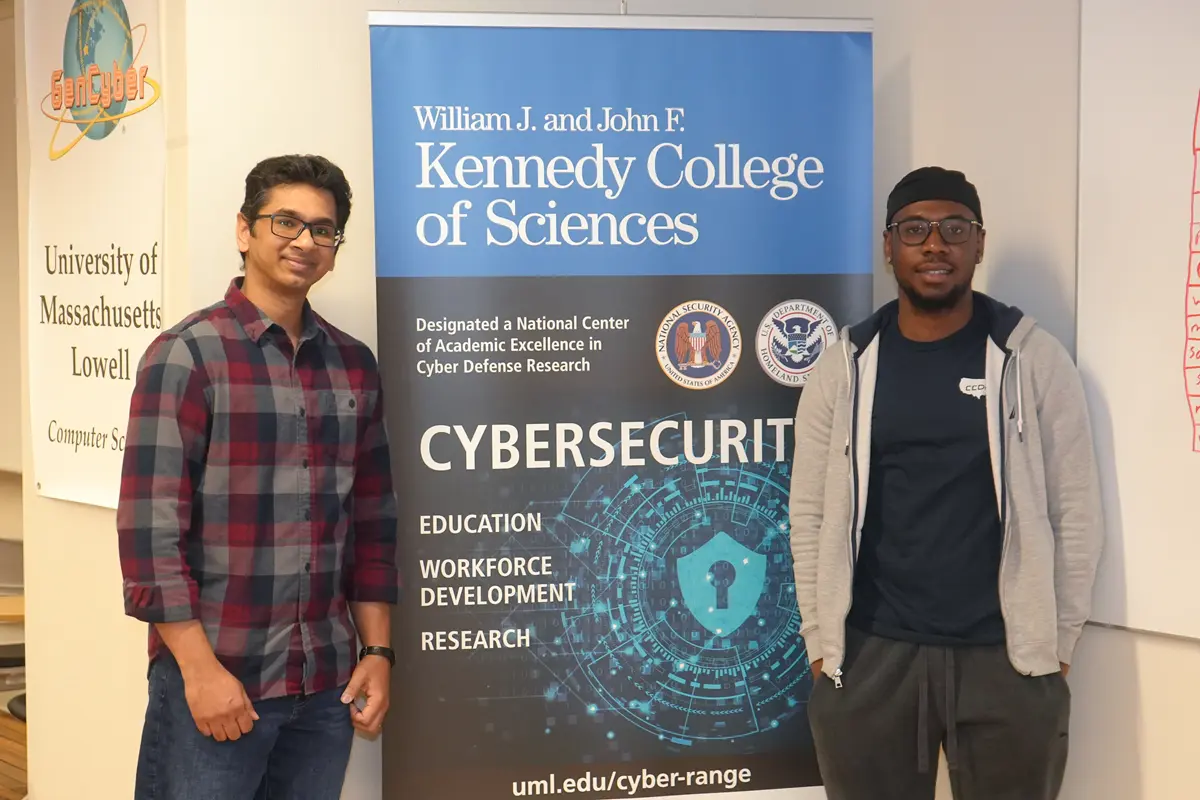 Two men pose for a photo while standing on either side of a sign for a cybersecurity range.