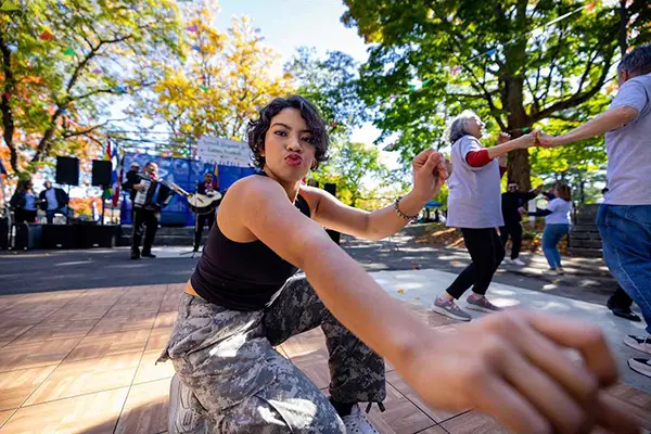 A woman with dark hair extends her arm to the camera while dancing on an outdoor stage