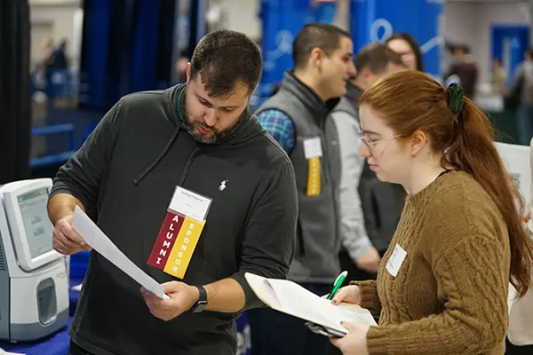 A man in a dark shirt looks over a resume with a young woman with glasses and long hair