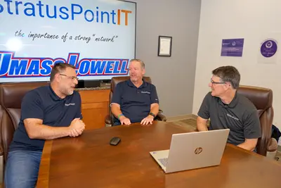 Three men in matching polo shirts talk while sitting around a laptop on a conference room table.