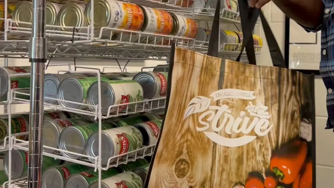UMass Lowell Strive grocery bag being held by hands in front of shelves loaded with canned goods.