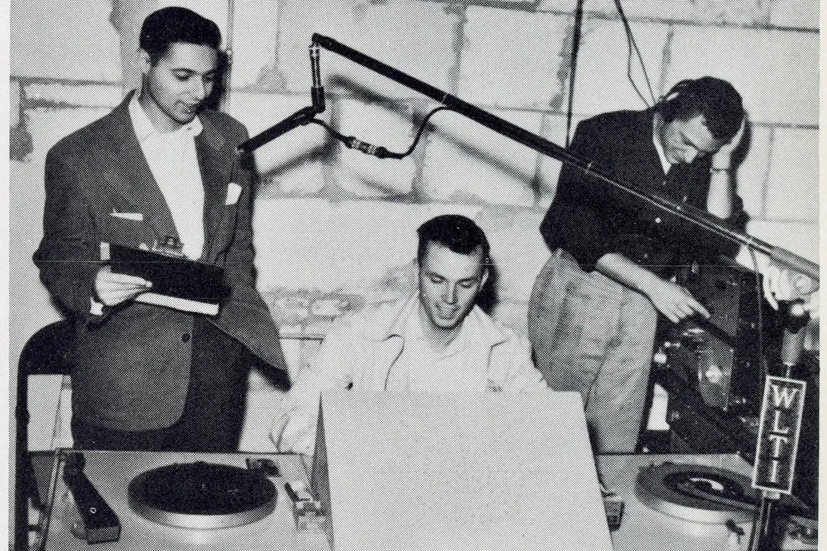 Three young men sit behind a radio station desk. On the desk are turntables and a microphone.