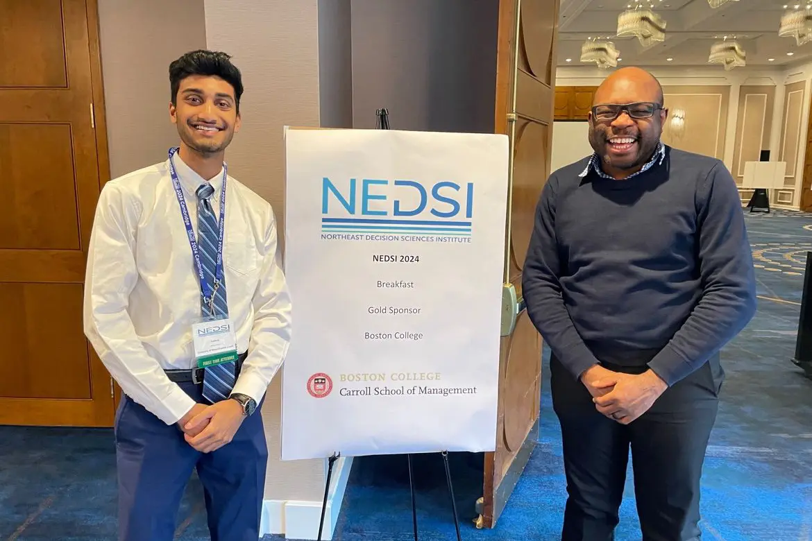 A college student and a professor pose for a photo while standing on either side of a research poster on an easel.