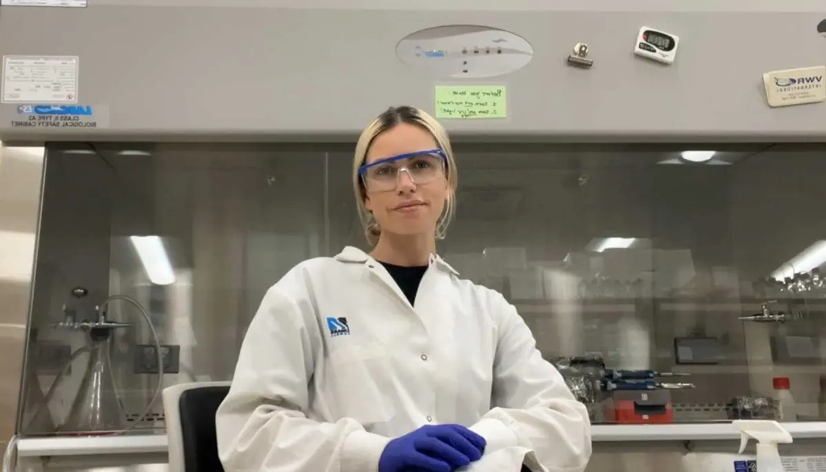   Sydney Sampson wears a lab coat and safety glasses in a UMass Lowell lab.