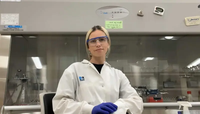   Sydney Sampson wears a lab coat and safety glasses in a UMass Lowell lab.