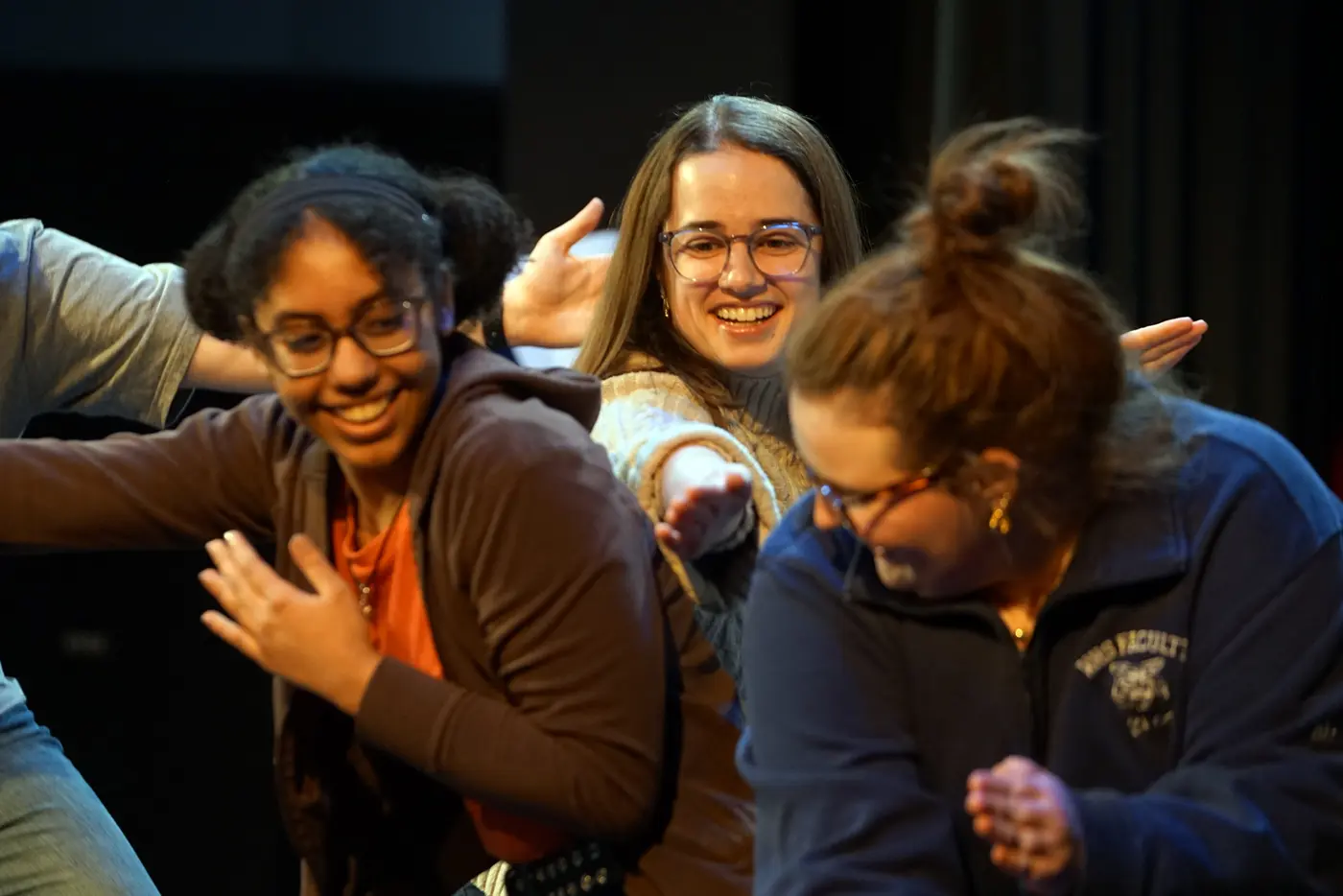 A smiling woman in glasses holds her arm between two younger women on a stage.