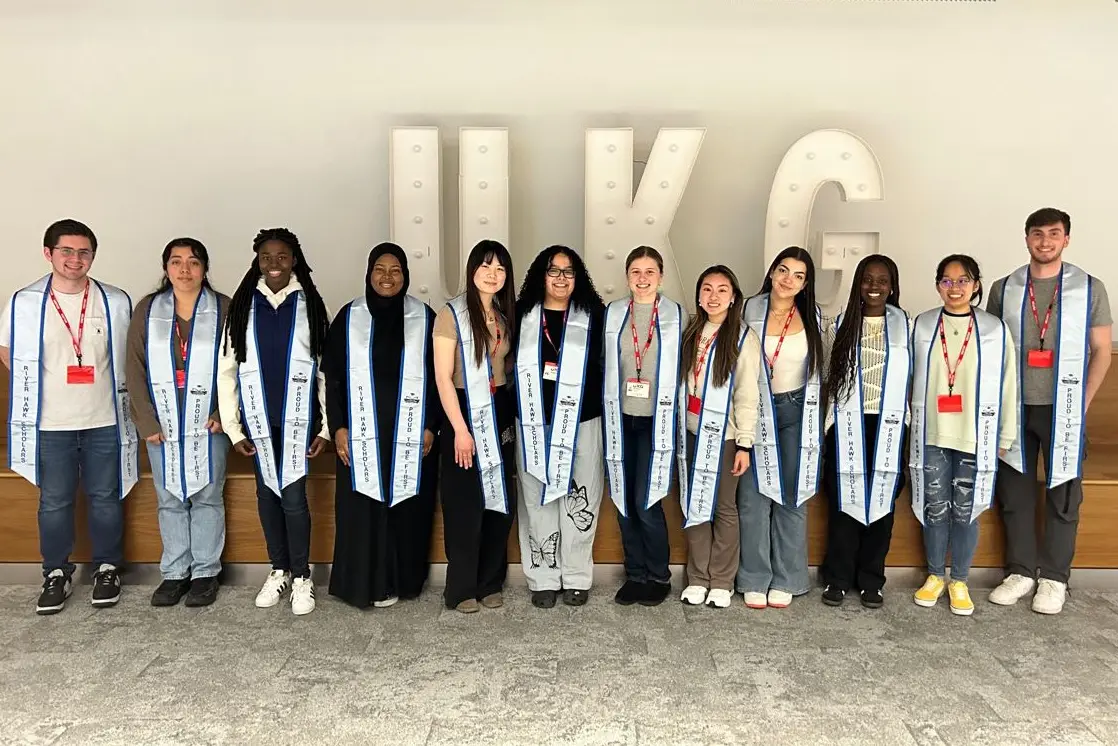 A dozen college students wearing stolls stand in a line and pose for a photo in front of a big UKG sign.