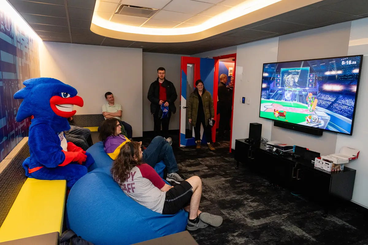 College students play a video game on a flatscreen TV while a college bird mascot looks on.