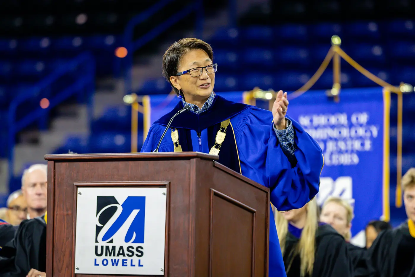 A woman wearing glasses and academic regalia gestures with her hand while speaking at a podium.