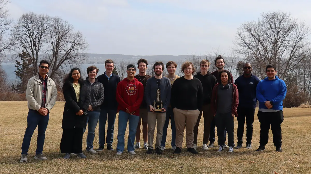 Fourteen people stand on a lawn in front of a bay.