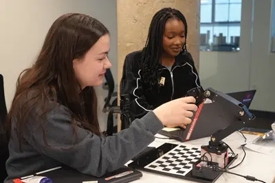 Vanessa Simoneau touches a robotic arm and Robina Nyangai types on a computer.