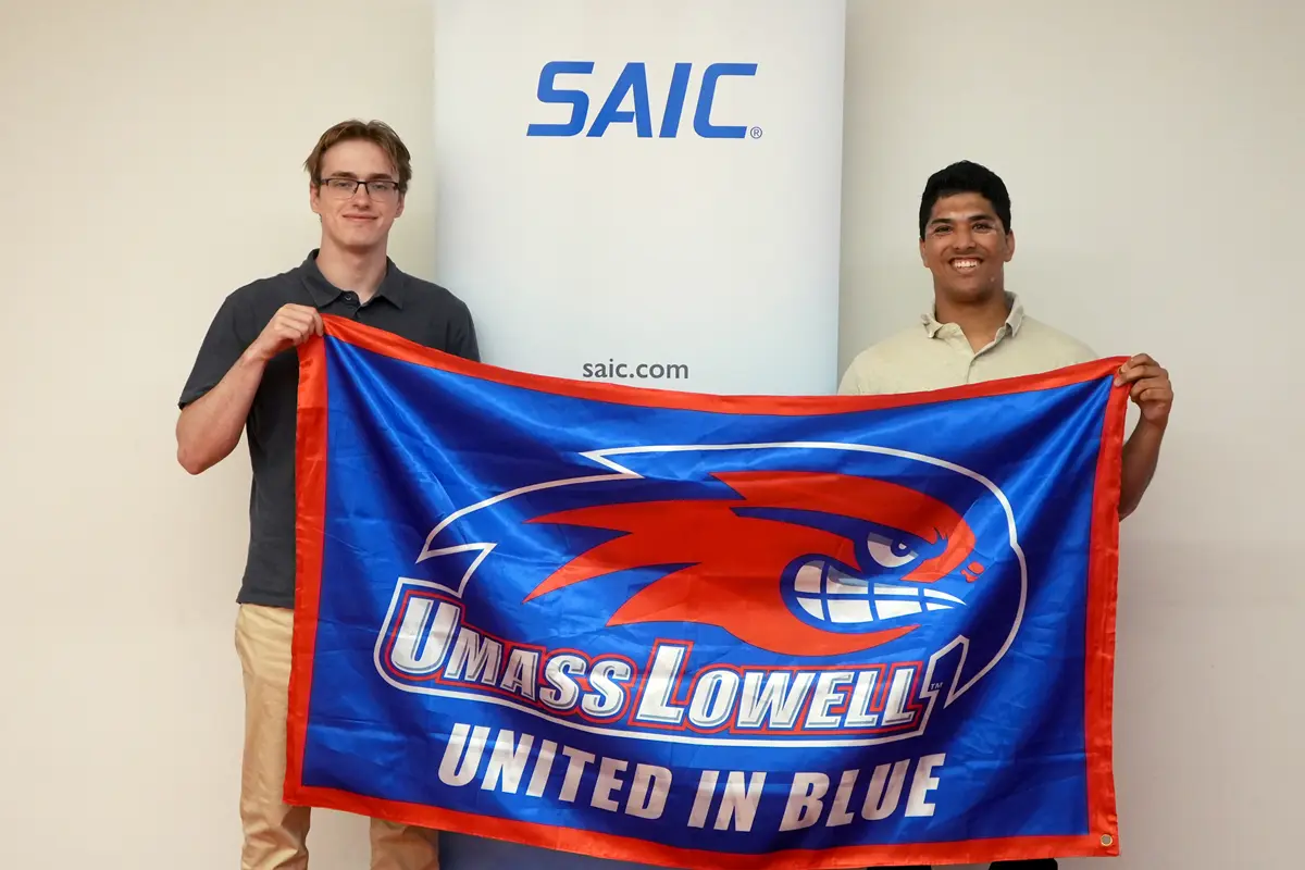 Two stuents hold a UMass Lowell flag in front of an SAIC banner.
