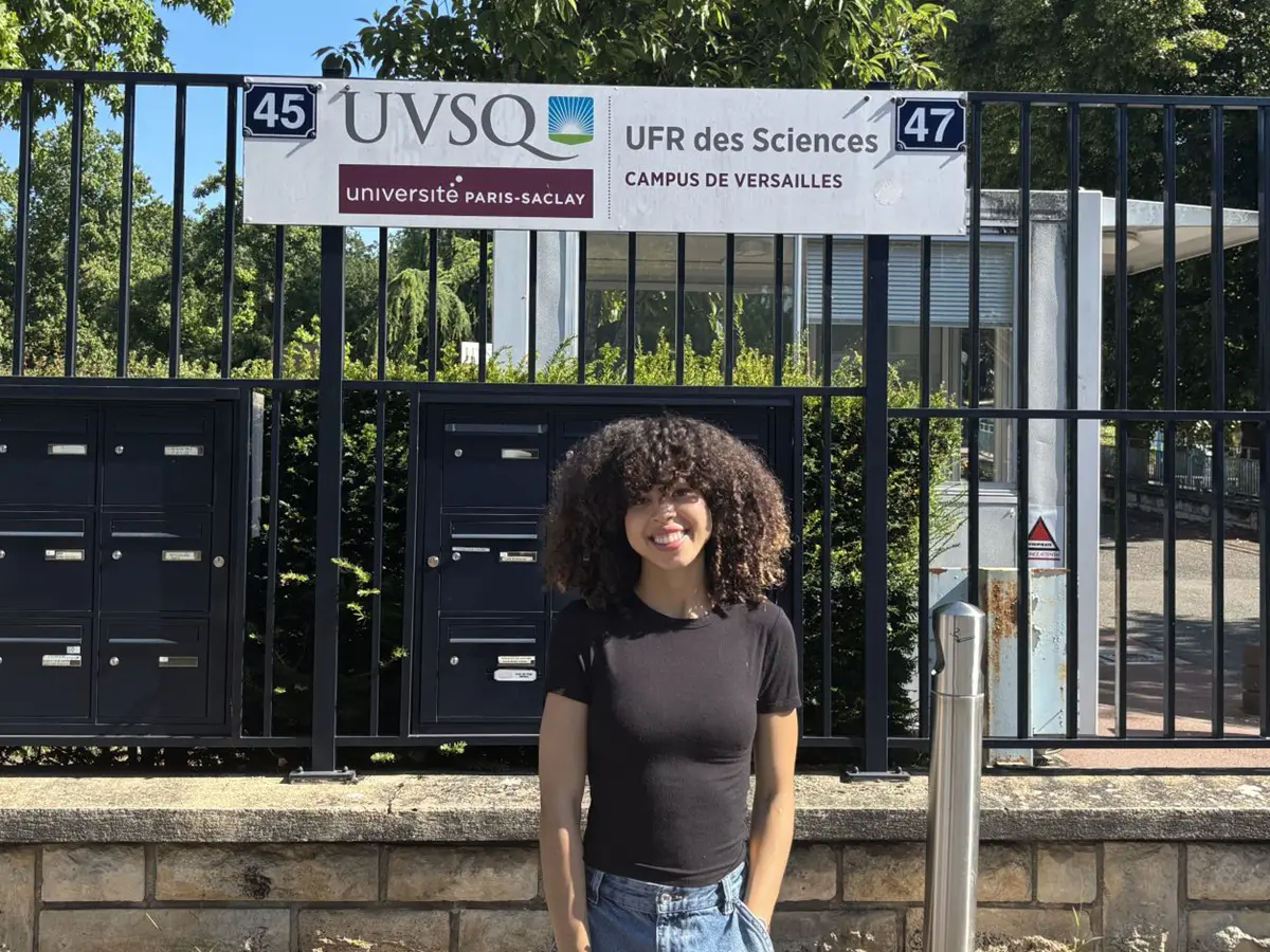 A woman in a black T-shirt poses for a photo outside in front of a fence with a sign.