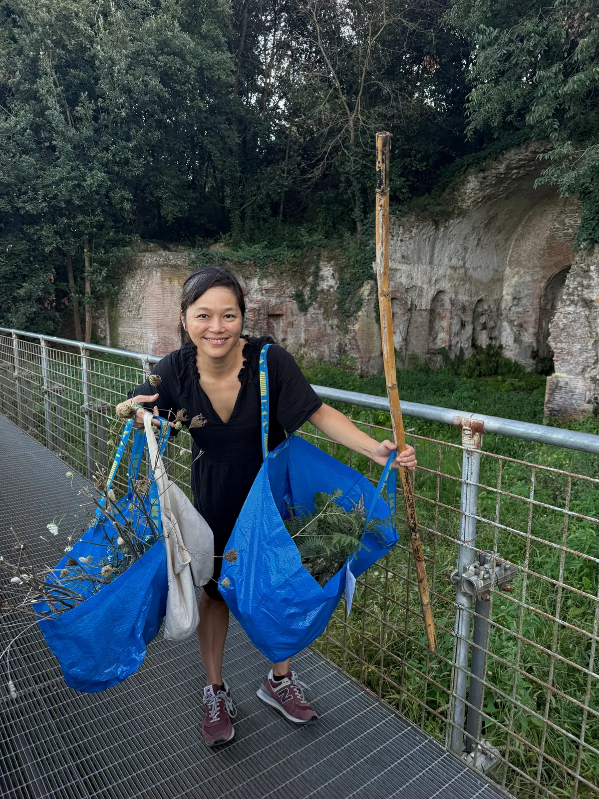 A woman carries two blue bags full of organic materials and holds a stick outside.