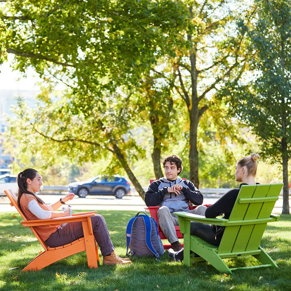 Three students sit on colorful Adirondack chairs under tree on UMass Lowell North Campus