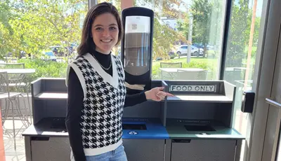 Student pointing to the food only slot of compost bin next to recycling bins.