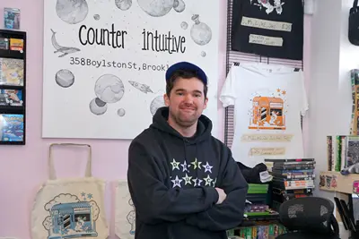 A young man in a black hoodie and baseball cap poses for a photo outside of a record store.