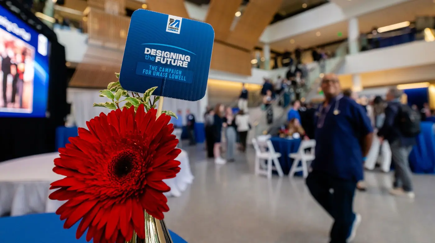 Designing the Future the campaign for UMass Lowell sign stuck in a bud vase with a red daisy in it while people have party in background
