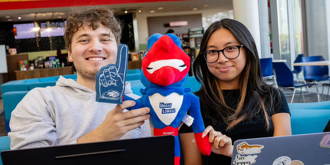 Two students hold a UMass Lowell Rowdy River Hawk doll holding a mini-No. 1 finger between them 