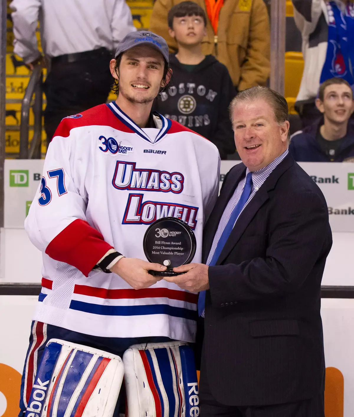 A college hockey goalie stands next to a man in a suit holding an award.