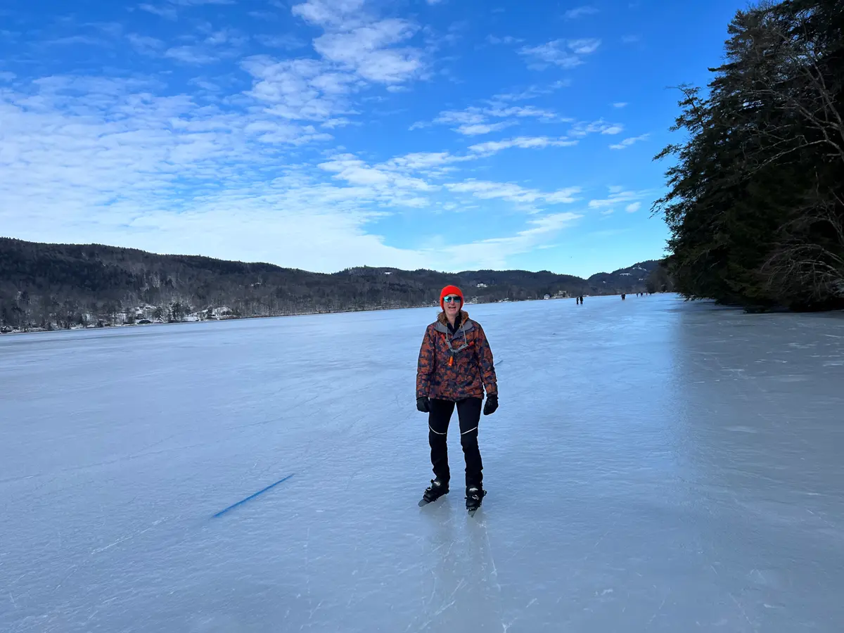 A woman in winter gear skates towards the camera on a totally smooth surface of lake ice with trees and sky behind.
