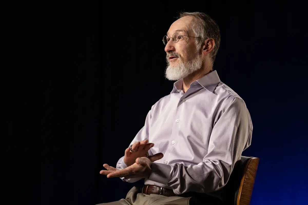 A man with a white beard and glasses gestures with his hands while sitting in a chair and talking.