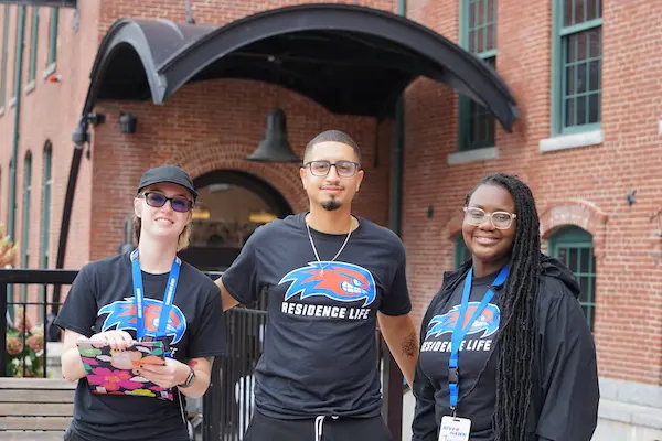 Three resident advisors wearing black UML T-shirts pose for a photo outside