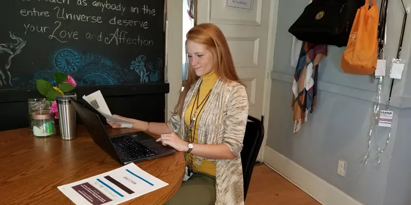 Nicole Haas sits at a table and works at her laptop at Revive Recovery Center in front of a decorated blackboard