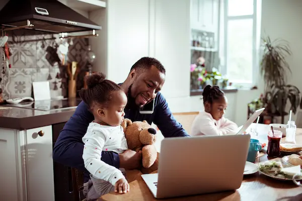A man talks on the phone and works on a laptop at home while holding his daughter on his lap