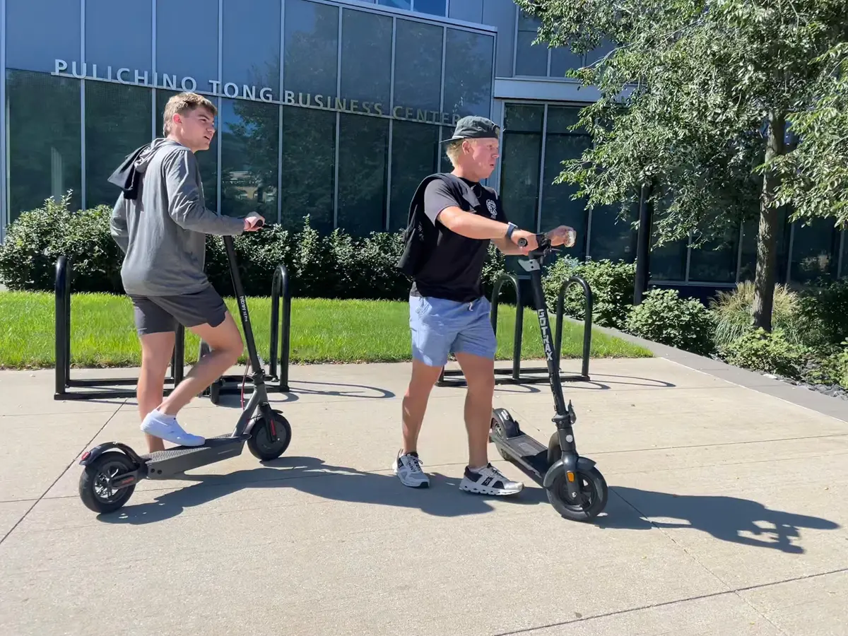 Two students prepare to ride electric scooters outside of a business school building.