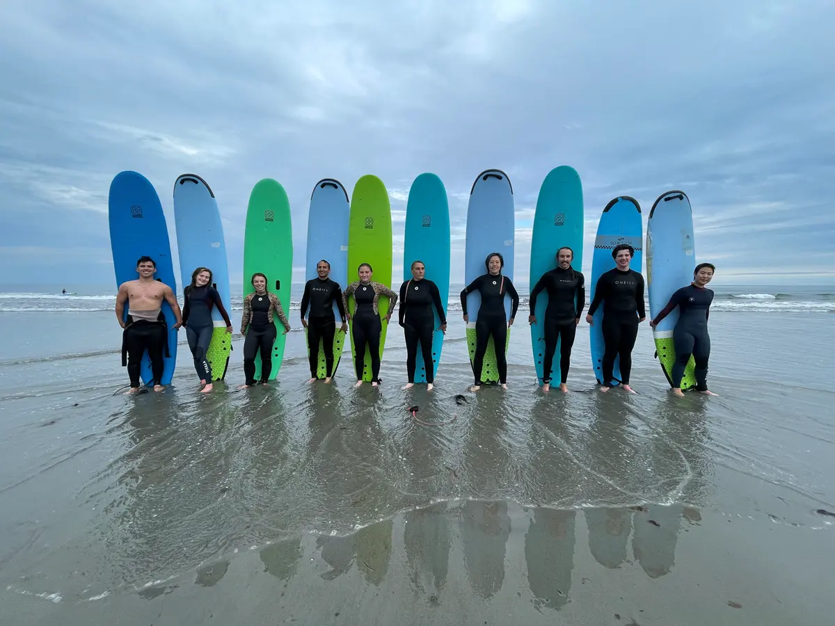 Ten people stand in a line holding surfboards behind them while standing on an ocean beach.