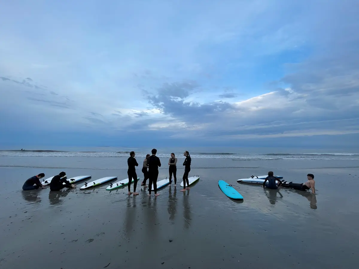 A group of surfers stand on the ocean shore. The sky is filled with clouds.