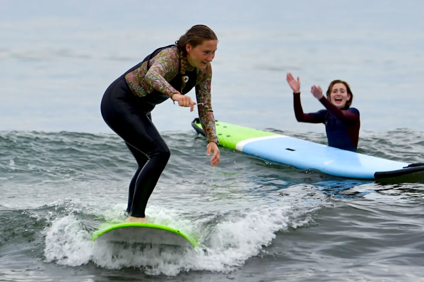 A person in a wetsuit stands on a surfboard while a person standing in the ocean behind them applauds.