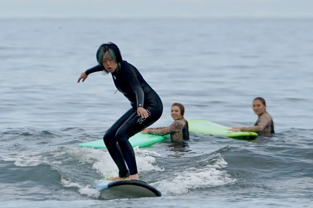 A person in a wetsuit rides a surfboard while two people in the ocean behind her look on.