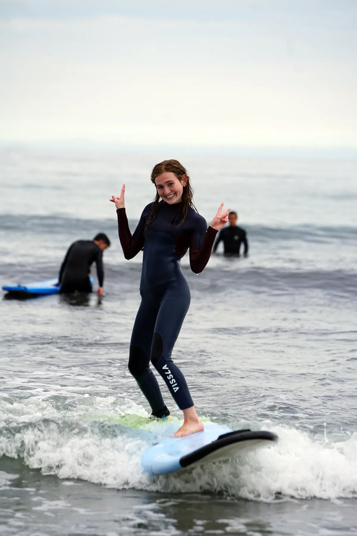 A woman in a wetsuit rides a wave on a surfboard while making peace signs with both hands.