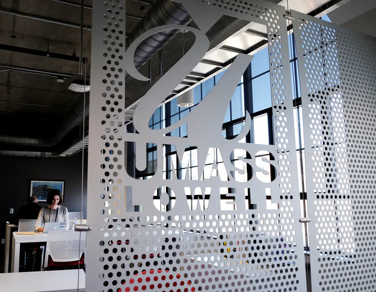 A UMass Lowell stencil/cut-out sign with a woman typing on a laptop computer in the background. Woman working at a desk behind a partition with the UMass Lowell logo.