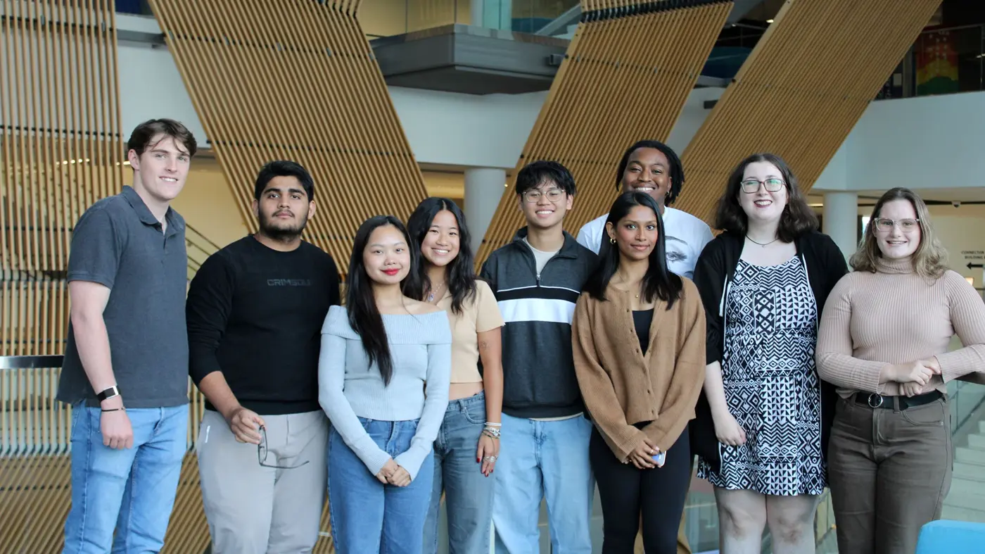 Nine students posing in front of wooden slat structure, smiling at camera